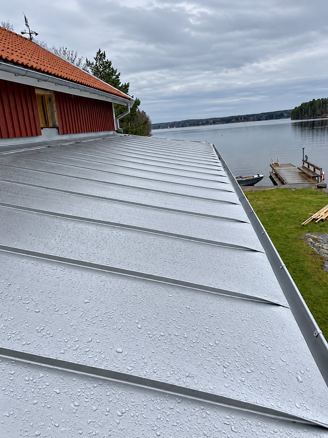 Ljusgrått falsat plåttak på hus vid vattnet, med brygga och båt i bakgrunden